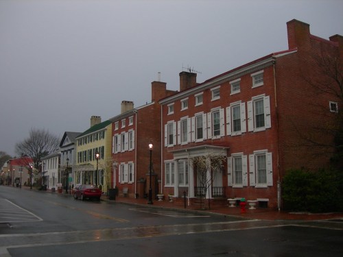 Historic brick buildings line a quiet street on a rainy day, with a red car parked nearby and streetlights glowing.
