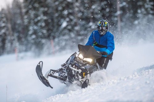 A person in a blue jacket rides a snowmobile through a snowy landscape, kicking up snow in the air.