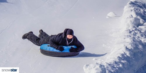 A person in winter gear is lying on a blue snow tube, sliding down a snowy slope.