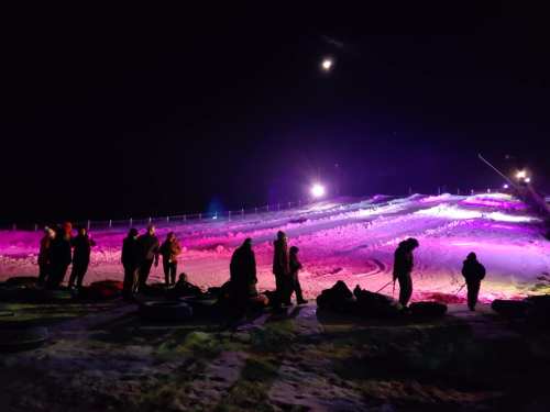 People gather on a snowy hill at night, illuminated by colorful lights and a bright moon.