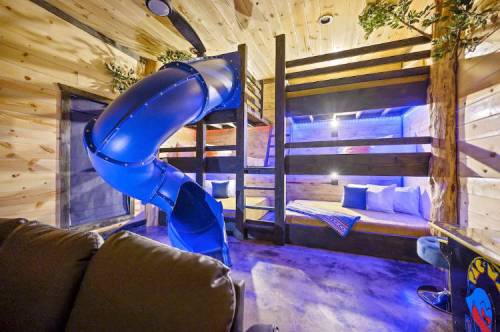 A cozy room with wooden walls, featuring a blue slide, bunk beds, and soft lighting for a playful atmosphere.