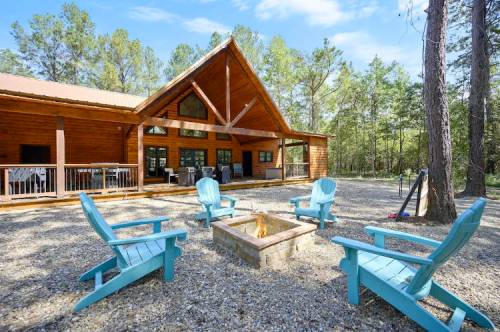 A cozy cabin surrounded by trees, featuring a fire pit and blue chairs in a serene outdoor setting.