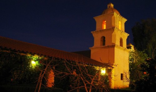 A historic building with a bell tower illuminated at night, surrounded by greenery and warm lights.