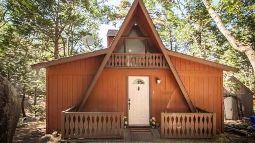 A charming A-frame cabin surrounded by trees, featuring a front porch and a welcoming entrance.