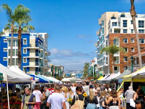 A bustling farmers market with crowds, tents, and palm trees, set against modern buildings and a clear blue sky.