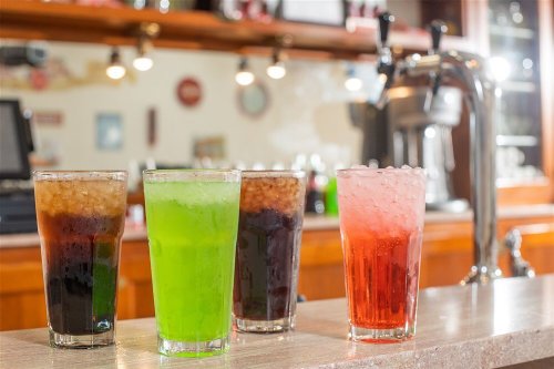 Four colorful drinks in tall glasses sit on a counter, with a bar in the background.