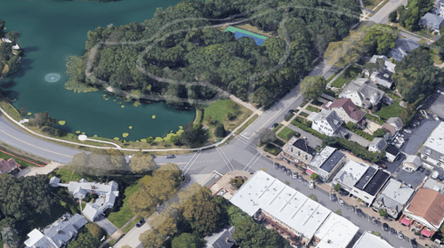 Aerial view of a park by a pond, surrounded by trees and residential buildings, with a road and shops nearby.