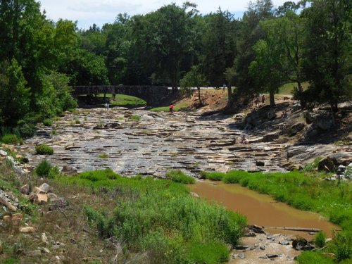 A rocky riverbed with patches of green grass and muddy water, surrounded by trees and a bridge in the background.