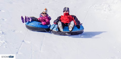 Two people in snow tubes sliding down a snowy hill, smiling and enjoying a winter day.