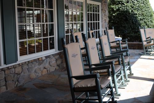 A row of rocking chairs on a stone patio in front of a building with large windows.