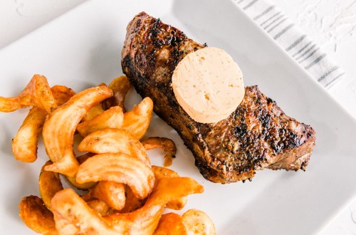 A grilled steak topped with a pat of butter, served with curly fries on a white plate.