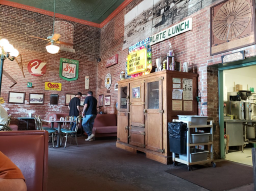 Interior of a vintage diner with brick walls, retro signs, and a wooden display cabinet. Two people walk in the background.