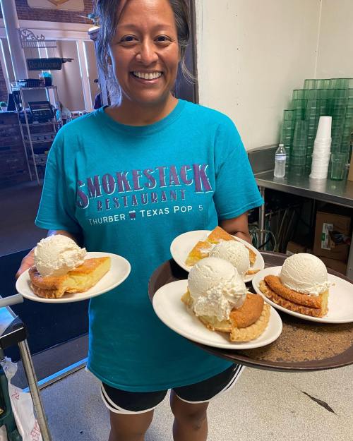 A smiling woman in a blue t-shirt holds four plates of dessert with ice cream in a restaurant setting.