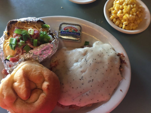 A plate with a baked potato topped with bacon and green onions, chicken smothered in gravy, a roll, and a side of corn.