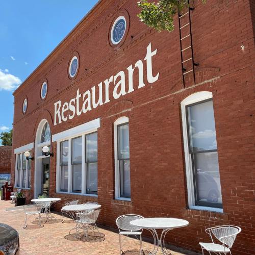 Exterior of a brick restaurant building with large windows and outdoor seating. Bright blue sky in the background.