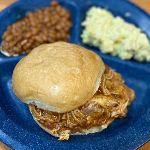 A plate with a pulled chicken sandwich, baked beans, and macaroni and cheese, all served on a blue tray.