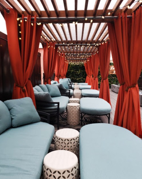 A cozy outdoor lounge area with teal seating, white tables, and red curtains, illuminated by string lights.