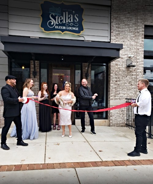 A group of people, including a woman in a dress, cut a red ribbon outside Stella's Elixir Lounge during a grand opening.