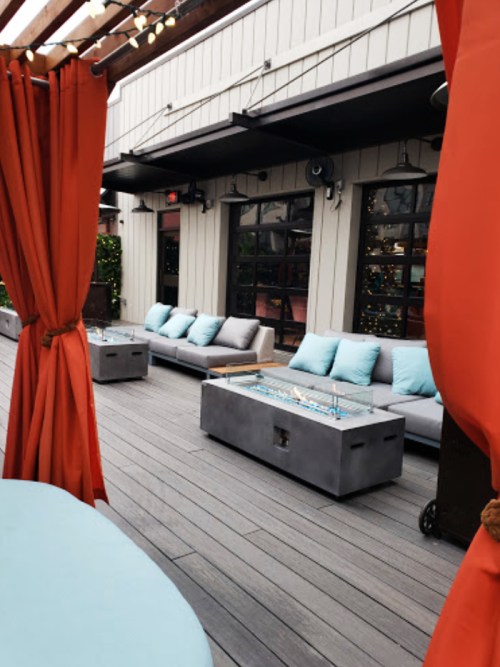 A rooftop lounge area with gray sofas, a fire pit, and orange curtains, surrounded by festive lighting.