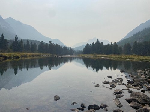 A serene river reflects mountains and trees under a hazy sky, with rocks lining the water's edge.