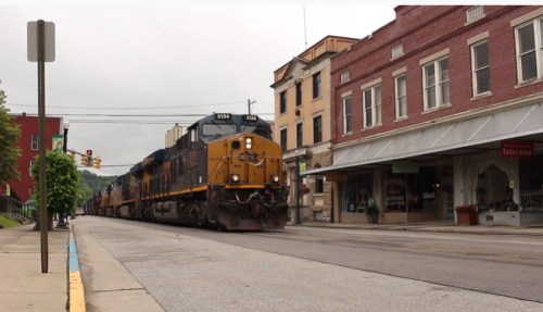 A train passes through a small town, with buildings lining the street and a cloudy sky overhead.