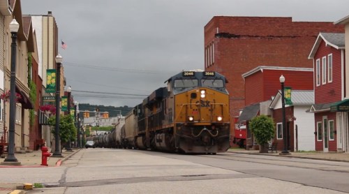 A freight train passes through a small town, with buildings and streetlights lining the road.