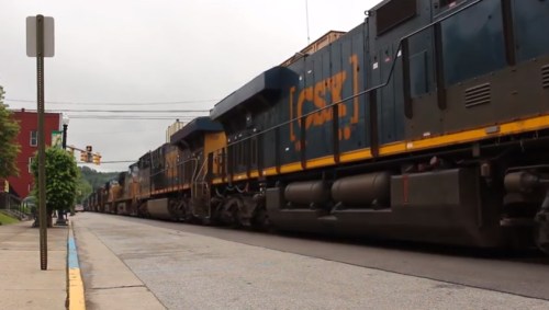 A freight train passes through a city street, with buildings and traffic signals in the background.