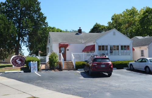 A white house with a red awning, sign for "Chocolate Storybook," and parked cars in a sunny parking lot.