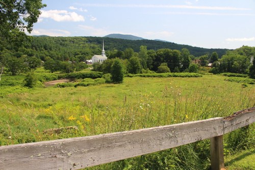 A scenic view of a green field with a wooden fence, mountains in the background, and a white church in the distance.