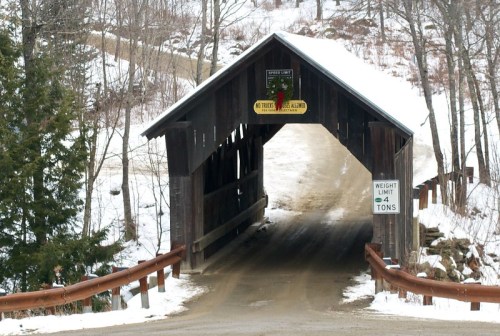 A snow-covered road leads through a wooden covered bridge, with a weight limit sign and festive decoration.