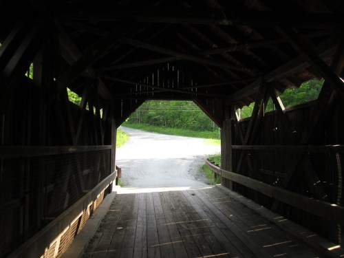 View from inside a covered bridge, looking out towards a winding road surrounded by greenery.