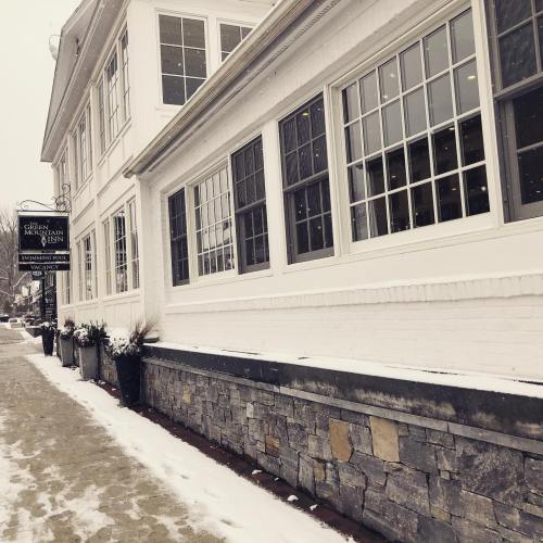 A snowy street scene featuring a white building with large windows and a stone wall, adorned with potted plants.