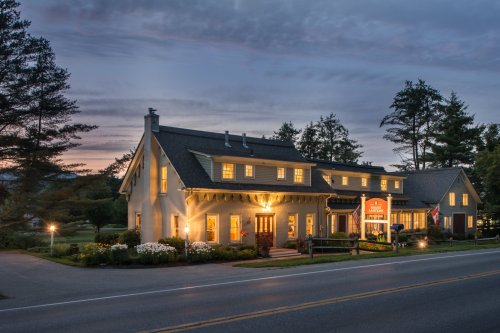 A charming two-story inn with warm lights, surrounded by trees, at dusk along a quiet road.