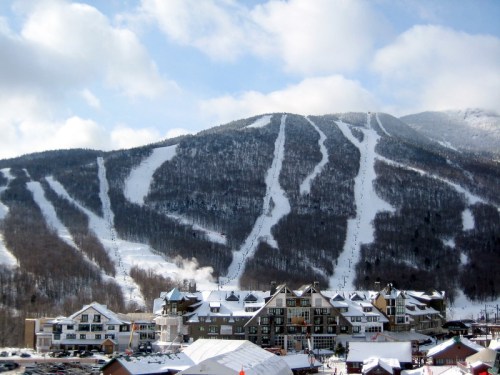 A snowy mountain with ski trails and a resort at its base under a partly cloudy sky.