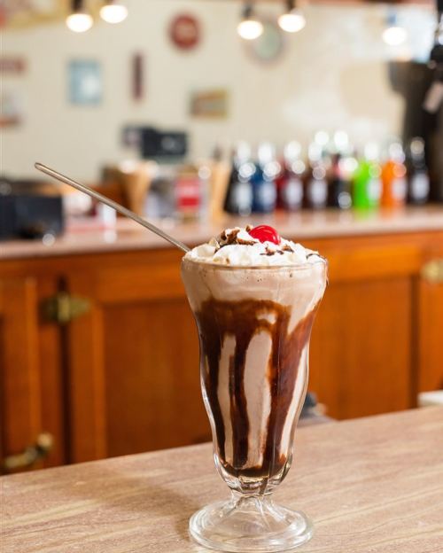 A tall glass of chocolate sundae topped with whipped cream and a cherry, set against a blurred background of a diner.