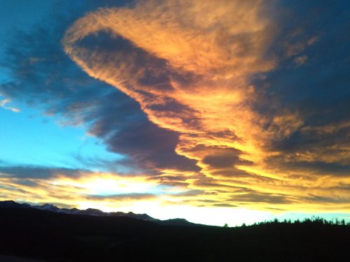 A vibrant sunset with swirling orange and yellow clouds against a blue sky, silhouetting distant mountains.