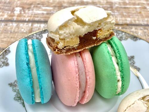 A plate with three colorful macarons: blue, pink, and green, with one macaron partially bitten into, revealing its filling.