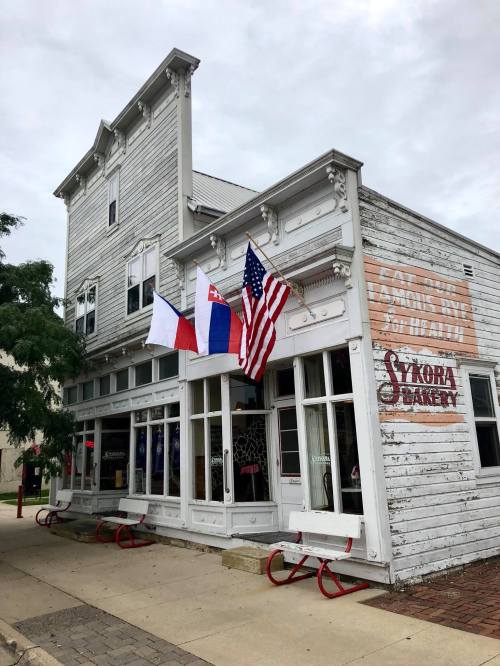A charming bakery with a white wooden exterior, featuring flags and a sign that reads "Sykora Bakery."