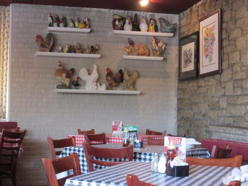 Cozy restaurant interior with checkered tablecloths, wooden chairs, and shelves displaying various decorative chickens.