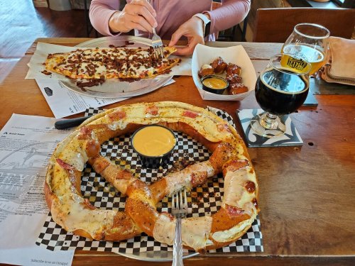 A large pretzel with cheese dip, a plate of loaded fries, and a glass of dark beer on a wooden table.
