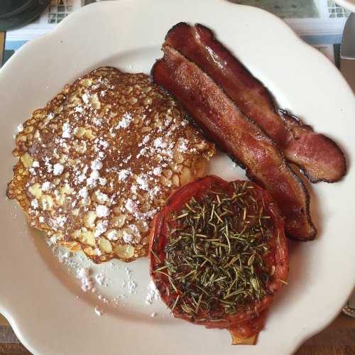 A plate with a fluffy pancake dusted with powdered sugar, crispy bacon, and a roasted tomato topped with herbs.