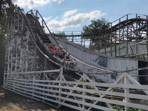 A group of people joyfully riding a wooden roller coaster on a sunny day, with a partially visible track and trees in the background.