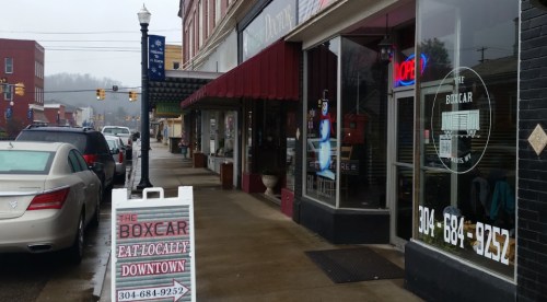 A street view of a small town with shops, a "The Boxcar" sign, and a parked car under a cloudy sky.