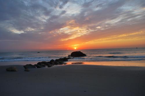 A serene beach at sunset, with vibrant orange and purple skies reflecting on the calm water and rocks along the shore.