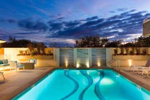 A serene pool area at dusk, featuring illuminated water, lounge chairs, and a colorful sky with clouds.