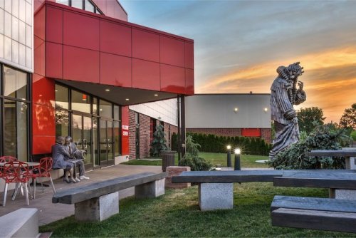 A modern building with red accents, featuring a large statue and outdoor seating area at sunset.