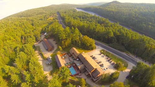 Aerial view of a lodge surrounded by dense forest and a winding river, with a pool and parking area visible.