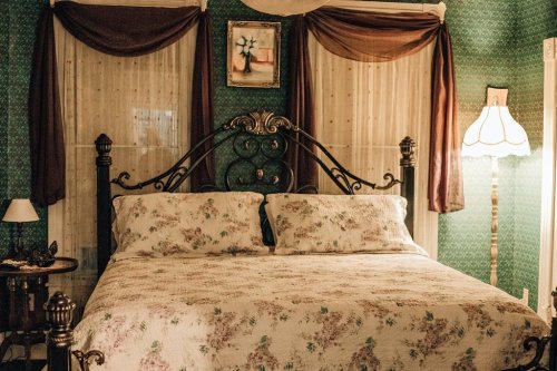 Cozy bedroom with a floral bedspread, ornate bed frame, vintage lamp, and patterned wallpaper.