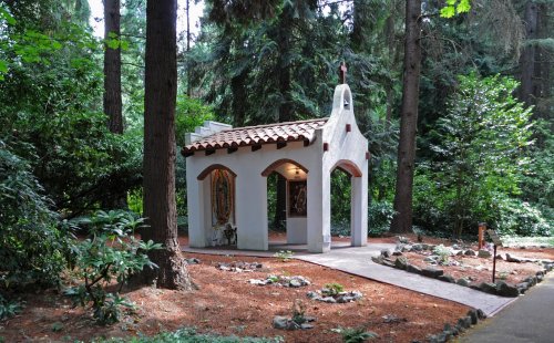 A small white chapel with a tiled roof nestled among tall trees and greenery in a serene forest setting.