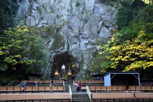 A natural rock formation with an open cave, surrounded by trees and empty wooden benches for an outdoor gathering.
